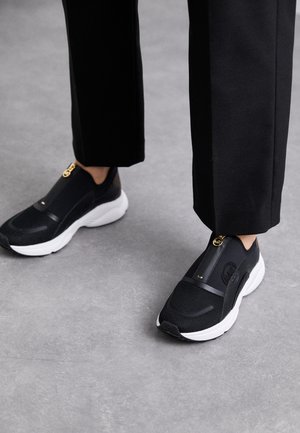 Black slip-on sneakers featuring a matte mesh upper, smooth accents, white sole, and gold hardware detail. Textured and sporty design.