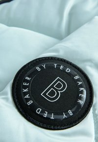 Svetlo modra satenska tkanina z okroglo črno oznako z logotipom, na kateri je bel "B", okoli pa napis "BAKER BY TED BAKER" v belem.