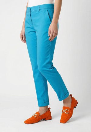 Osoba nosząca jasnoniebieskie, dopasowane spodnie ze podwiniętymi nogawkami oraz pomarańczowe mokasyny z złotym łańcuszkiem, stojąca na białym tle.