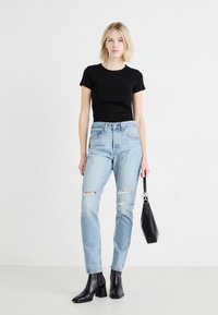 Zwarte T-shirt met korte mouwen, lichtblauwe distressed jeans, zwarte hakschoenen tot de enkel en een kleine zwarte tas, tegen een neutrale achtergrond.