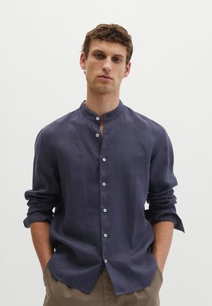 Junger Mann mit Händen in den Taschen, trägt ein marineblaues Hemd mit Mandarinkragen und hochgekrempelten Ärmeln bis zu den Ellbogen.