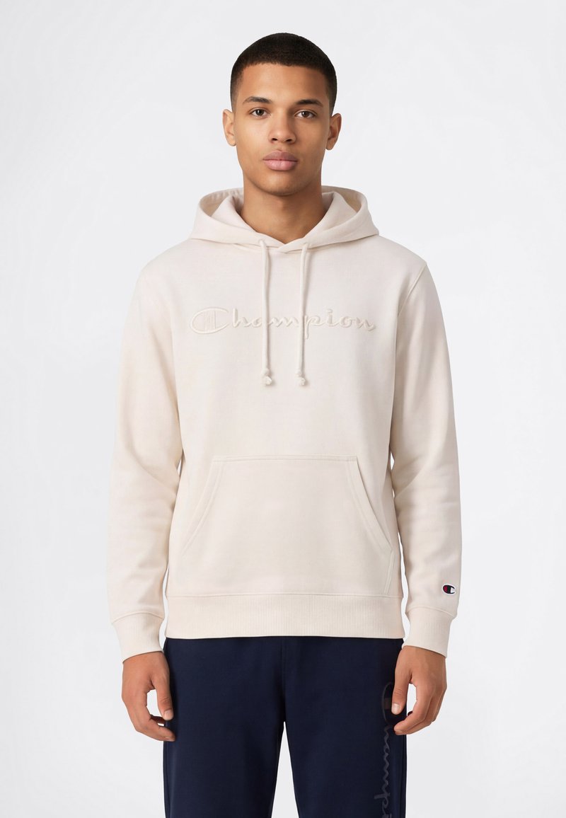 Sweat à capuche couleur crème avec logo "Champion" brodé, cordons de serrage, poche kangourou et poignets côtelés. Associé à un pantalon foncé.