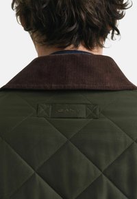 Gequilte groene jas met een bruin corduroy kraag en een label waarop "GANT" staat. De textuur is glad, met diamantvormige stiksels.