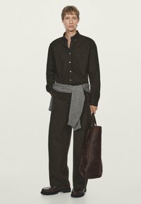 Giovane uomo con camicia button-up marrone scuro e pantaloni, maglione grigio annodato in vita, che tiene una grande borsa a tracolla in pelle marrone scuro.