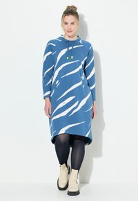 Hooded blauwe jurk met witte abstracte strepen, gemaakt van zachte stof, met lange mouwen en een relaxte pasvorm, in combinatie met beige enkellaarsjes.