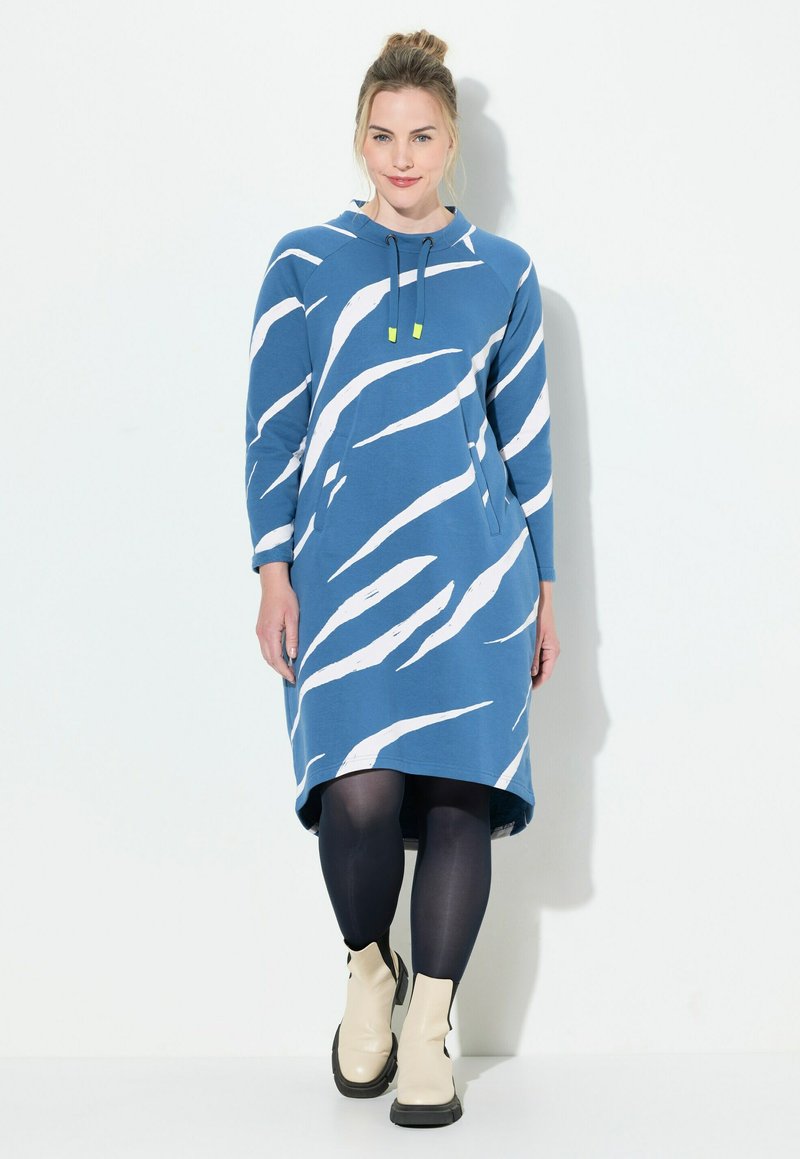 Hooded blauwe jurk met witte abstracte strepen, gemaakt van zachte stof, met lange mouwen en een relaxte pasvorm, in combinatie met beige enkellaarsjes.
