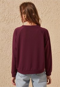 Sudadera burdeos con mangas raglán largas, cuello redondo y un corte relajado, combinada con jeans azul claro frente a una pared texturizada.
