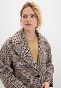 Brun och blårutig kappa med stora slag, enstaka knappstängning och överdimensionerade ärmar. Buren över en beige rundhalsad tröja.