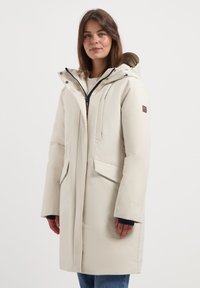 Jonge vrouw in een lichte beige winterjas met capuchon, met rits en flapzakken, staand tegen een effen witte achtergrond.