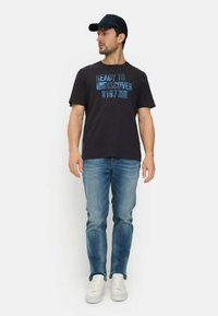 Sort T-shirt med blåt print "READY TO DISCOVER 1977", båret med lyseblå jeans og hvide sneakers, suppleret med en mørk kasket.