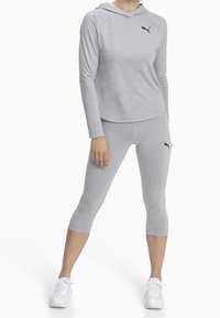 Haut de sport gris avec capuche et manches longues, associé à un legging capri gris. Les deux pièces présentent un accent logo noir. Baskets blanches.