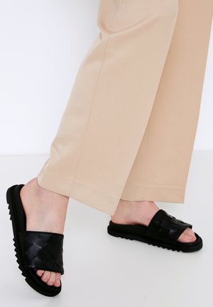 Piernas con pantalones beige y sandalias tipo diapositiva negras tejidas con suelas gruesas sobre un fondo blanco.
