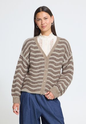 Femme portant un cardigan beige et marron à rayures ondulées par-dessus un chemisier en dentelle blanche et un pantalon bleu, debout devant un fond uni.
