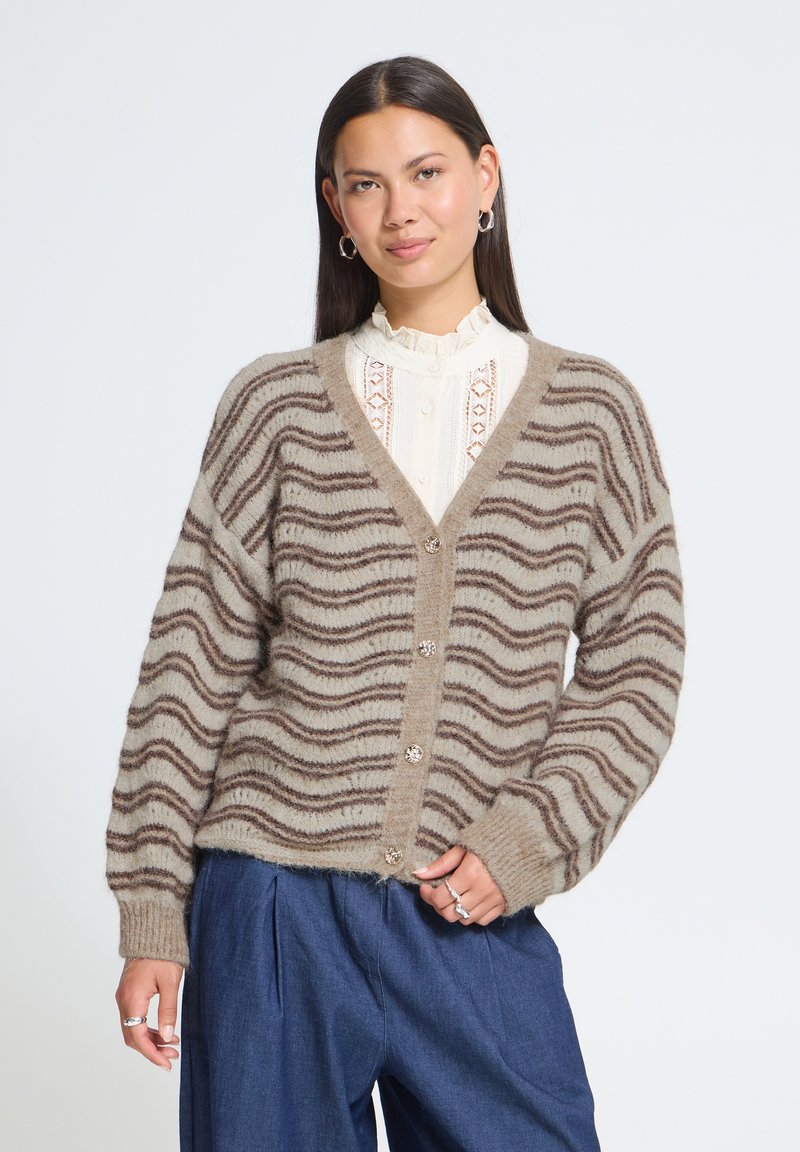Femme portant un cardigan beige et marron à rayures ondulées par-dessus un chemisier en dentelle blanche et un pantalon bleu, debout devant un fond uni.