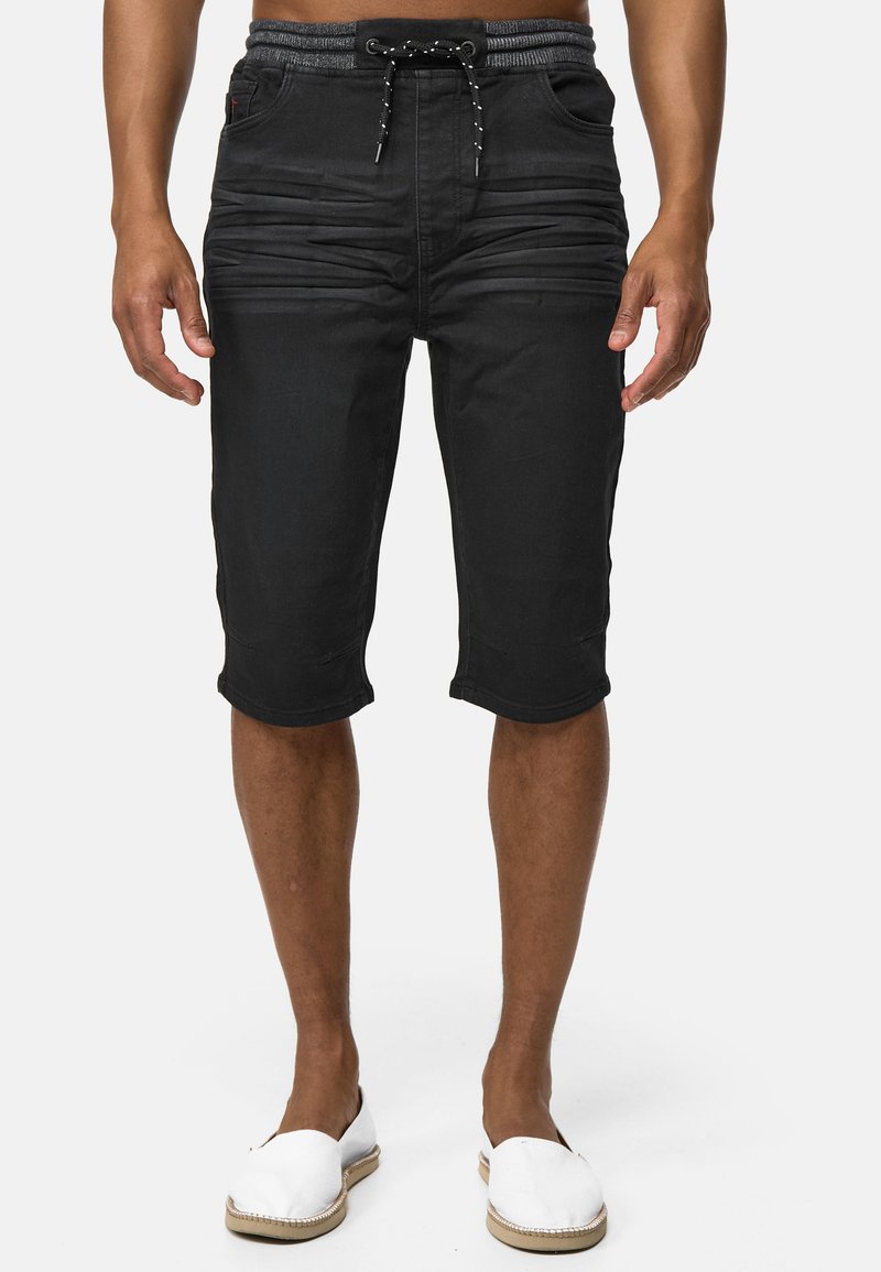 Pantalones cortos de mezclilla negra con cintura ajustable, detalles texturizados en la parte frontal y dobladillos enrollados. Combinados con zapatos slip-on blancos.