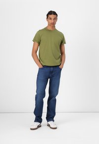 Zeleno bombažno majico s zavitimi rukavi je kombinirana z modrimi jeans hlačami. Manekenka stoji z rokama v žepih in nosi bele superge.