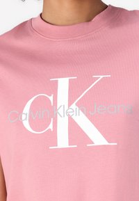 Rosa bomull t-skjorte med stor hvit "K" og grå "Calvin Klein Jeans" tekst, med rund hals og korte ermer.