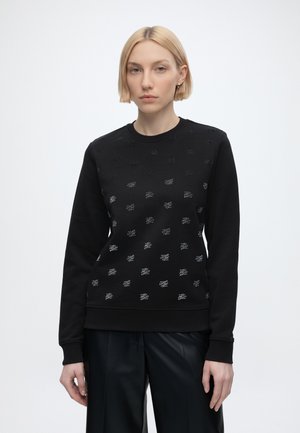 Donna con capelli biondi corti che indossa un maglione nero a maniche lunghe con piccoli loghi bianchi decorati e pantaloni neri su uno sfondo semplice.