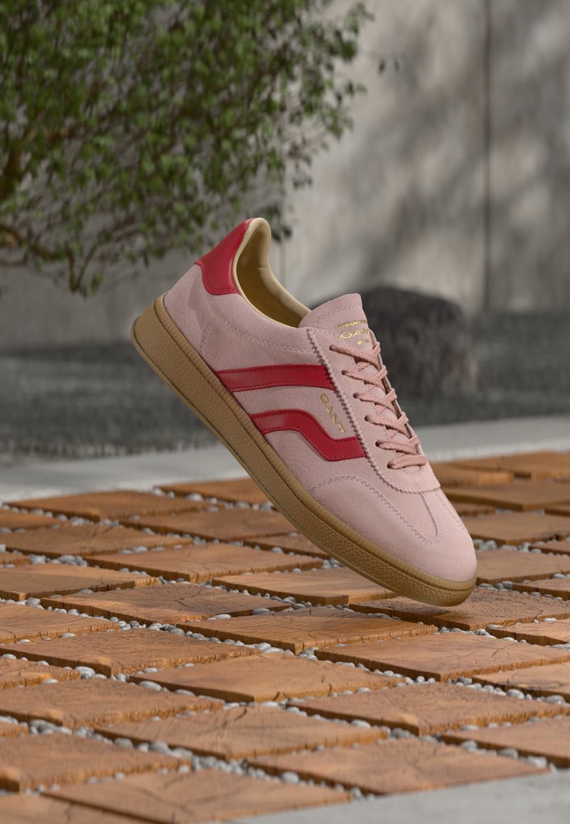 Sneaker in camoscio rosa con accenti rossi e marchio dorato. Presenta un design con lacci e suola in gomma, mostrata sopra una superficie di legno texturizzata.