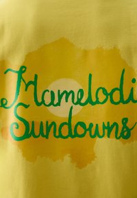 Camiseta amarilla con el texto "Mamelodi Sundowns" en verde, con un gráfico de sol en el fondo y una suave textura de acuarela.
