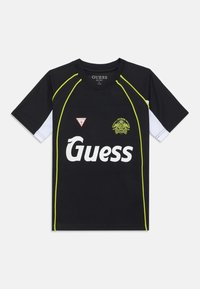 Svart idrettsdrakt med korte hvite ermer, neon grønne detaljer og en stor hvit "Guess"-logo. Har et trekantet merke på venstre side.