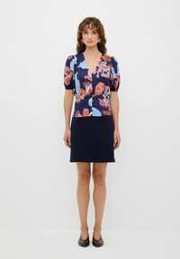 Blumenbluse mit Puffärmeln in Marineblau, Orange und hellblauen Mustern. Kombiniert mit einem figurbetonten Marineblau-Rock und braunen Schuhen.