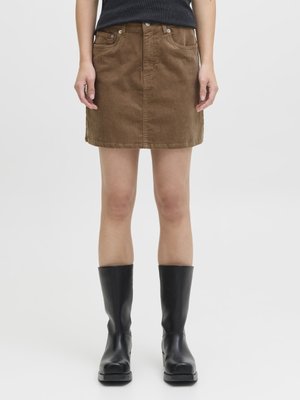 Άτομο που φορά καφέ μίνι φούστα από κορντόρι, μαύρο αμάνικο τοπ και μαύρες δερμάτινες μπότες μέχρι τη μέση της γάμπας, στέκεται σε απλό φόντο.