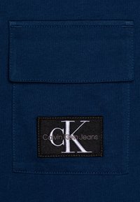 Mörkblå bomullstyg med bröstficka. Har en svart etikett med "CK" och "Calvin Klein Jeans" i silverbroderi.