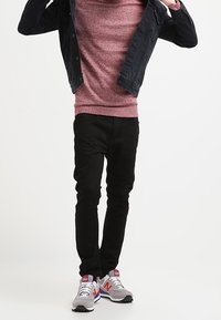 Avslappnad outfit med en mörk denimjacka, röd mönstrad tröja, svarta slim-fit byxor och grå sneakers med rosa detaljer.