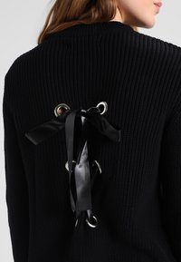 Zwarte gebreide trui met een geribbelde structuur, voorzien van grote zilveren oogjes en zwarte satijnen linten die door de achterkant zijn geregen voor verstelbare stijl.