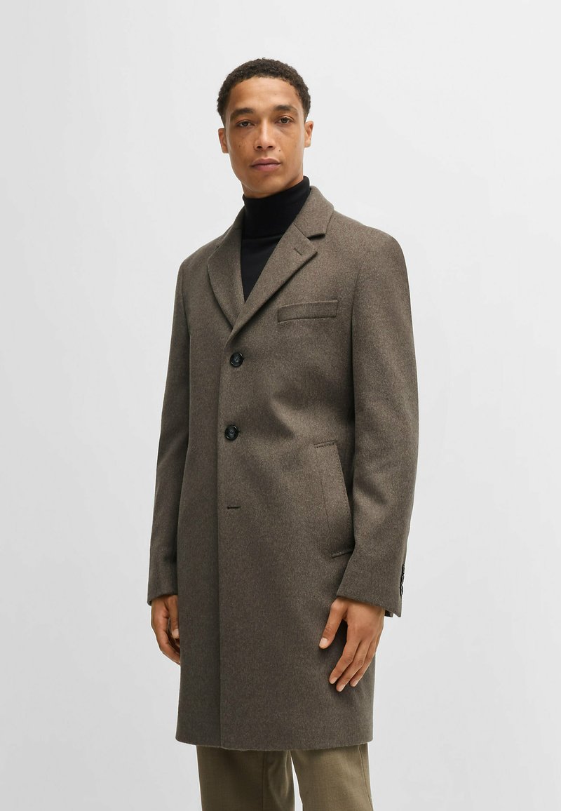 Cappotto di lana marrone con colletto a scollo, due bottoni e tasca sul petto. Presenta un taglio dritto e tasche laterali. Indossato sopra un dolcevita nero.