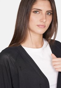 Cardigan nero in maglia con scollatura a V, caratterizzato da dettagli testurizzati e piccoli accenti di perline. Indossato sopra una maglietta bianca. Texture liscia.