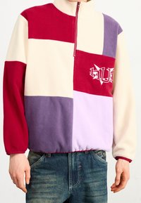 Fleecepullover med høj krave, som har skiftende blokke af rød, creme, lilla og lavendel. Inkluderer lynlås-lukning og logo-detalje.