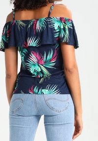 Kwiecisty top z odkrytymi ramionami w kolorze granatowym, z żywymi zielonymi i różowymi wzorami liści, z falbanką przy dekolcie. W komplecie z jasnoniebieskimi dżinsami.