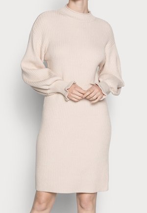 Femme portant une robe pull beige en maille côtelée avec de longues manches ballon et un col montant, debout devant un fond uni.