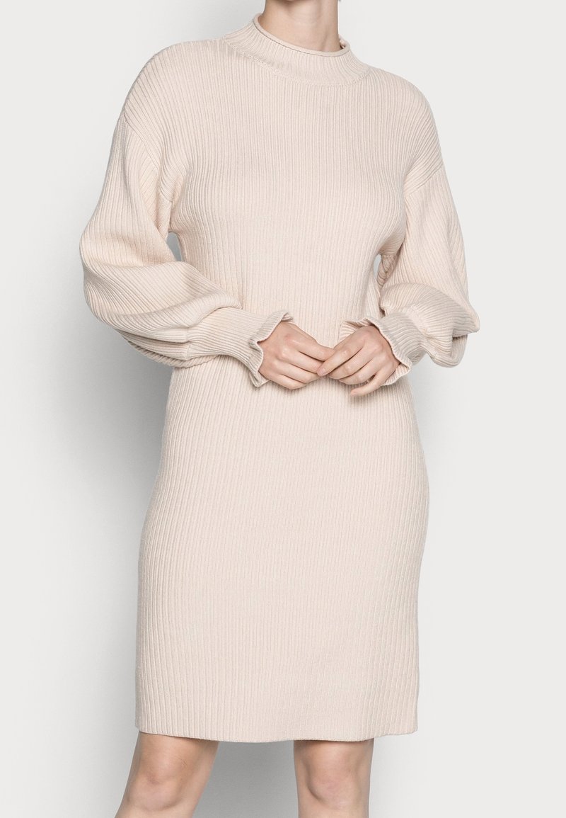 Femme portant une robe pull beige en maille côtelée avec de longues manches ballon et un col montant, debout devant un fond uni.