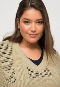 Beige Strickpullover mit V-Ausschnitt und offener Webstruktur an den Schultern. Über einem dunklen Hemd getragen, das ein strukturiertes Muster aufweist.