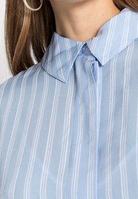 Camicia azzurra chiaro con colletto a righe verticali bianche. Il tessuto appare liscio e leggermente lucido. Presenta una chiusura con bottoni e un colletto strutturato.