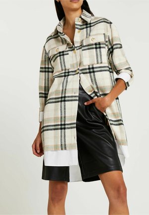 Mujer vestida con una camisa a cuadros beige de botones dorados sobre una camisa blanca, acompañada de pantalones cortos de cuero negros hasta la rodilla.