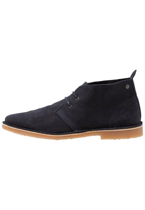 Bottine en daim noir avec deux œillets, bout rond, semelle en caoutchouc beige et languette au talon, vue de profil sur fond blanc.