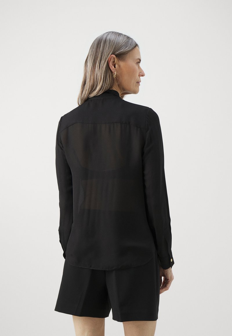 Czarna bluzka z długim rękawem i przezroczystym tyłem, dopasowany krój i mankiety zapinane na guziki. Miękka tkanina i zaokrąglony dół, w zestawie z dopasowanymi szortami.