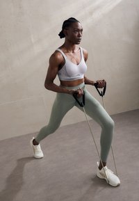 Sports-bh i lys grå med svarte detaljer, høy midje mintfargede leggings og hvite treningssko. Modellen bruker motstandsbånd i en treningsposering.