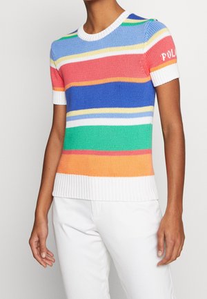 Persoon draagt een gebreide trui met korte mouwen en ronde hals, met horizontale regenboogstrepen en een witte geribde zoom, gecombineerd met een witte broek.