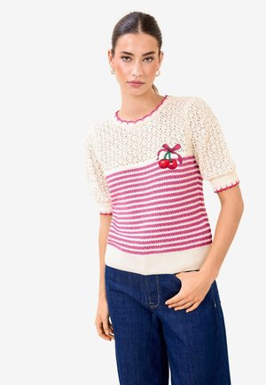Mladá žena v krémovo-ružovom pruhovanom pletenom tričku s výšivkou čerešní a tmavomodrých džínsoch pózuje s jednou rukou v vrecku.