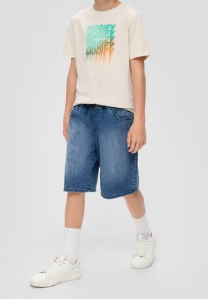 Camiseta de algodón beige con texto gráfico en teal y naranja, combinada con pantalones cortos de mezclilla azules y zapatillas blancas con cordones visibles y calcetines.