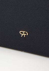 Mörkblå textilyta med subtil textur, med en liten guldfärgad logotyp framtill som framhäver kontrast och varumärkesdetaljer.