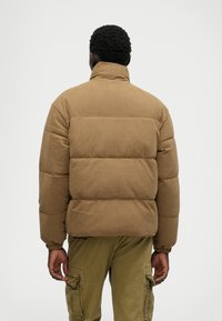 Bruine corduroy pufferjack met een hoge kraag, gewatteerd ontwerp en elastische manchetten. Draag het over olijfgroene cargo-broeken, gezien van achteren.