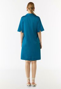 Teal Kurzarmkleid mit Kragen, lockerer Passform, mittellanger Schnitt bis zur Oberschenkelmitte und Seitenventilation, kombiniert mit geschnürten Sandalen mit Absatz.