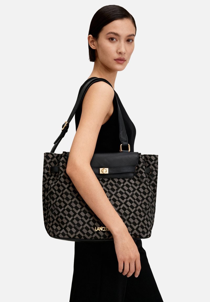 Borsa tote con motivo geometrico nero e grigio, con patta in pelle nera e dettagli dorati, dotata di due manici robusti e una forma strutturata.