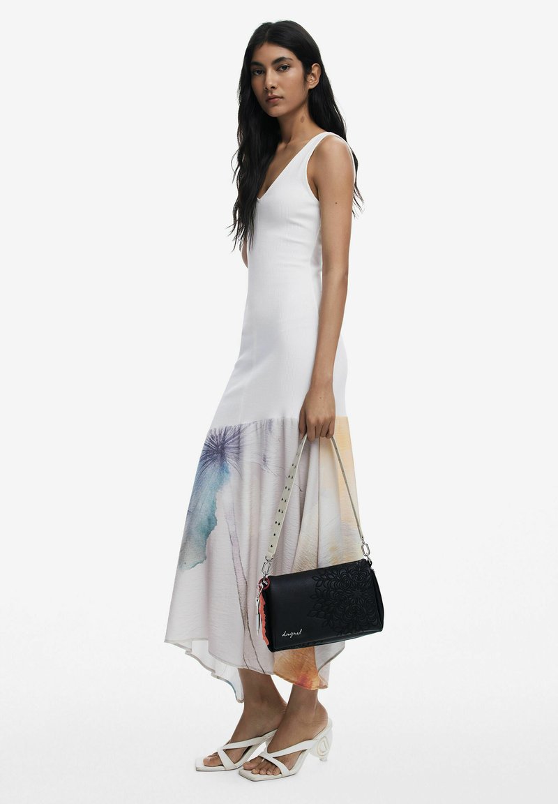 Borsa a spalla nera con motivo floreale in rilievo, accessori in argento e una tracolla a contrasto. Indossatrice in un vestito bianco senza maniche con bordo colorato.